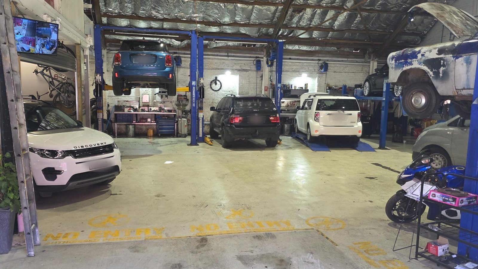 The Harold Park Repairs workshop seen from the roller-door entrance: cars on hoists either side of the drive-through bay, with workbenches and tooling along the wall