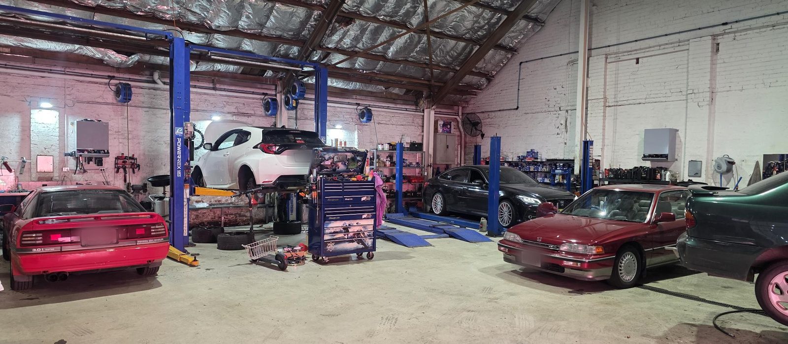 Inside the Harold Park Repairs workshop: a car raised on a hoist under the timber roof trusses, with more cars, tool chests and parts shelving along the painted brick walls